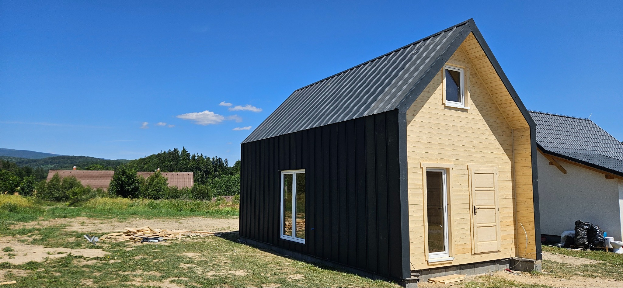 Nowoczesny dom z dachem z blachy trapezowej i elewacją z desek w kolorze naturalnego drewna i czarnym. Widok z boku na tle zieleni i błękitnego nieba.