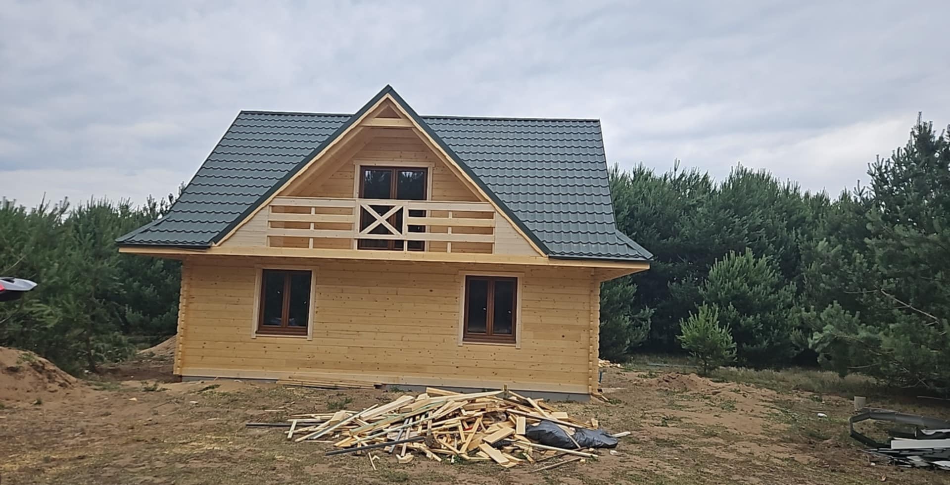 Drewniany dom z ciemnoszarym dachem z blachodachówki, widok z przodu. Na pierwszym planie sterta drewna i czarna folia. W tle las iglasty i pochmurne niebo.