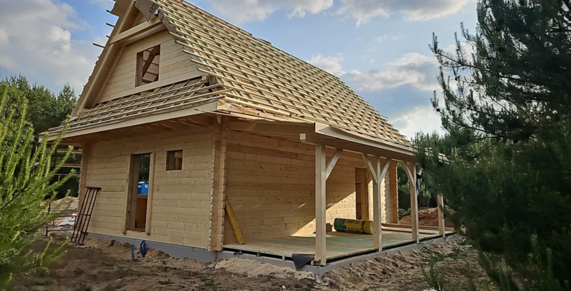 Drewniany dom w trakcie budowy, widoczne elementy konstrukcji dachu i werandy. Na pierwszym planie iglaste drzewo, w tle błękitne niebo z chmurami.