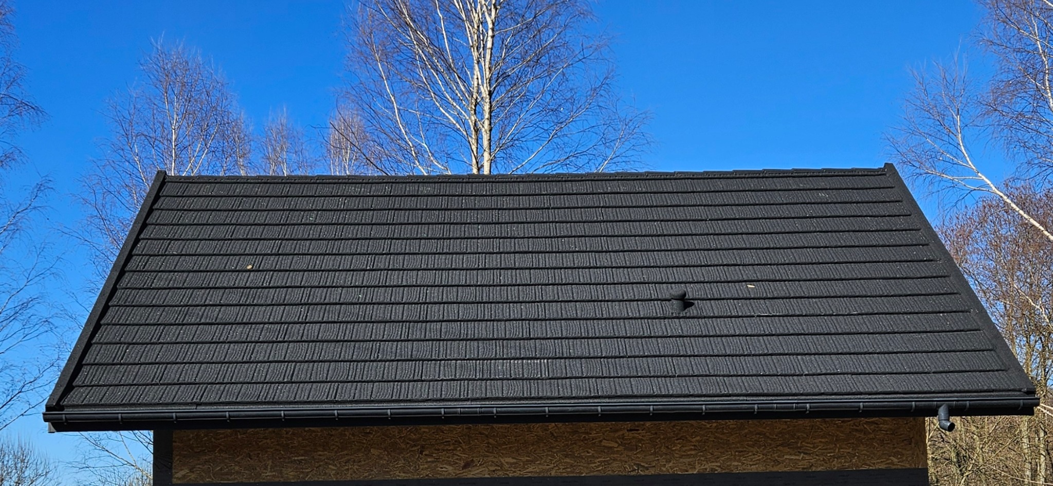 Czarny dach z blachodachówki modułowej, z widoczną rynną i drzewami w tle, prezentowany w słoneczny dzień. Detal pokrycia dachowego.