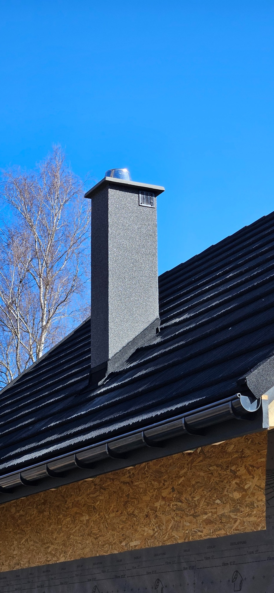 Grafitowy dach z blachodachówki modułowej, szara rynna i komin z nasadą, na tle błękitnego nieba i drzewa bez liści. Widoczna surowa ściana szczytowa.