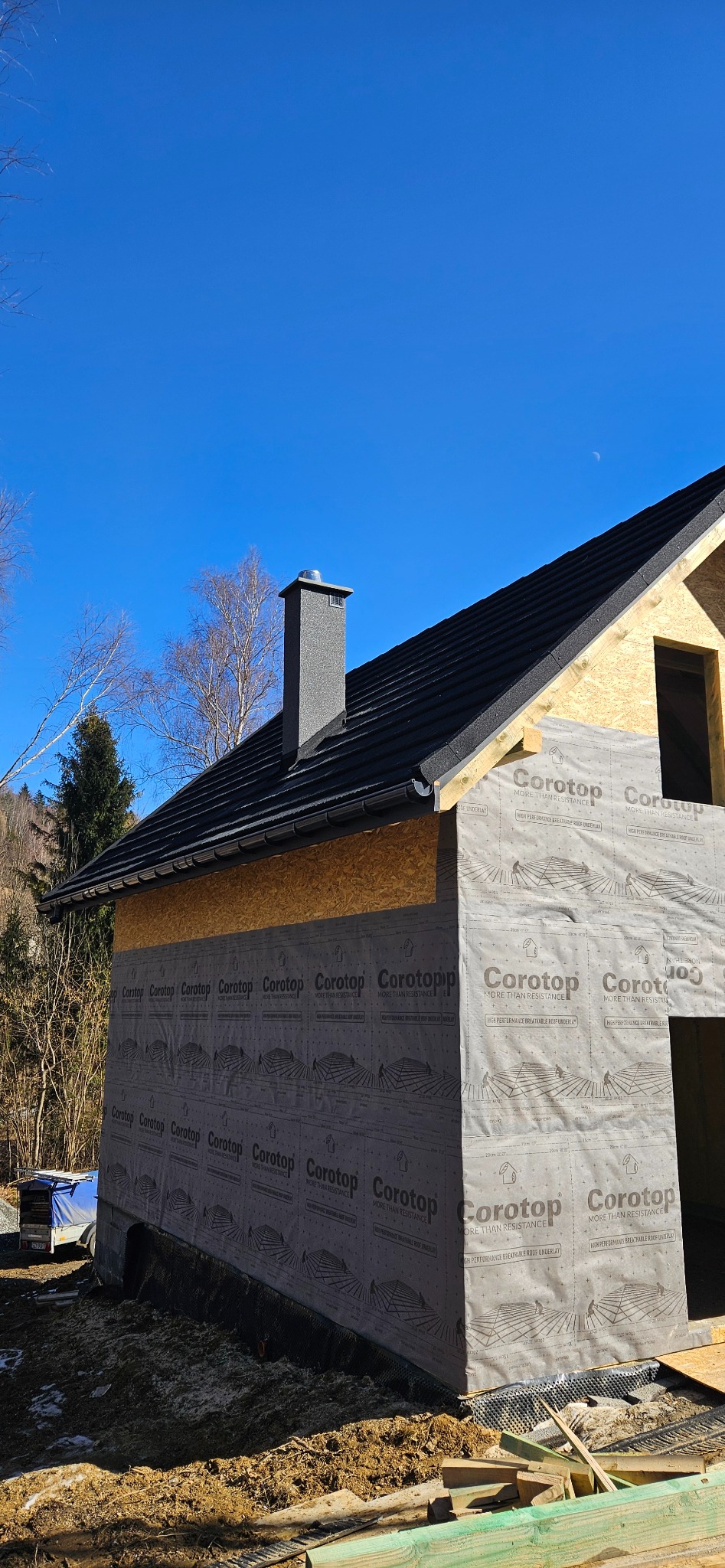 Dach z kominem i membraną Corotop na budynku w Jeleśni, w trakcie prac. Widoczne deski i elementy konstrukcyjne. Błękitne niebo w tle.