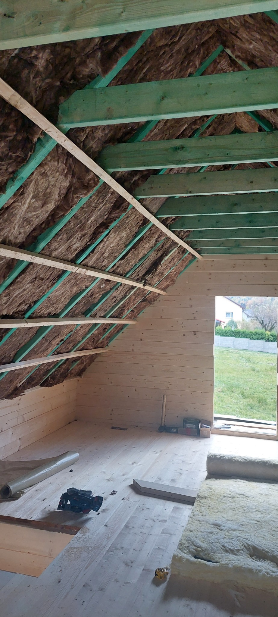 Izolacja poddasza wełną mineralną, widoczne drewniane elementy konstrukcyjne dachu i ściany z desek. Na podłodze rozłożona wełna i narzędzia.