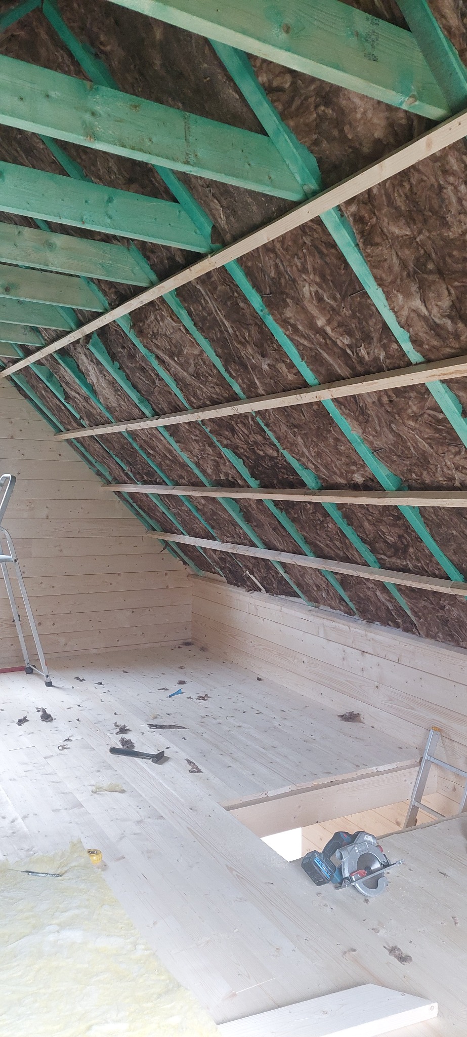 Poddasze w trakcie izolacji: widoczne drewniane krokwie, wełna mineralna między nimi i deski podłogowe. Na podłodze leży piła elektryczna.