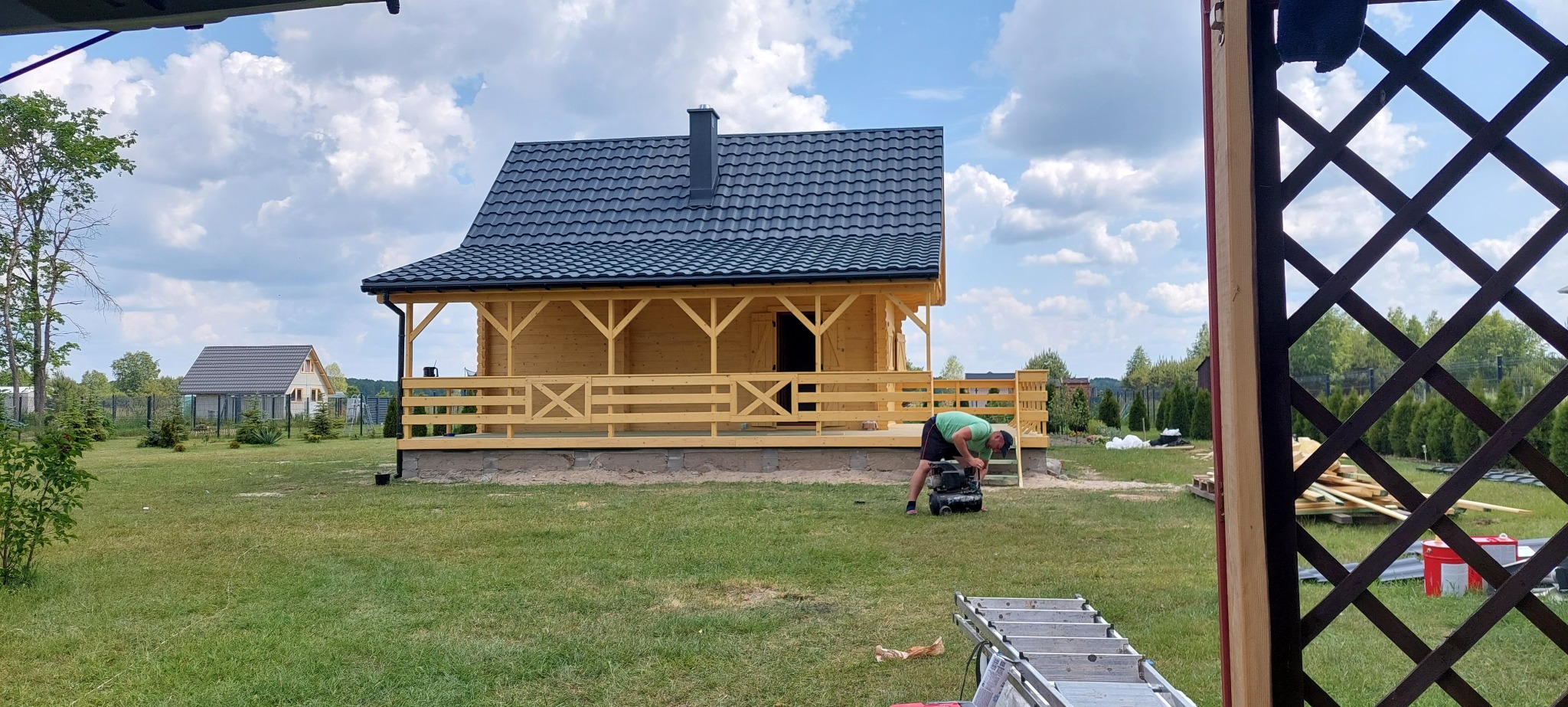 Drewniany dom z ciemnym dachem z blachy trapezowej, widok z zewnątrz, na pierwszym planie mężczyzna przy kompresorze, drabina. W tle drugi dom i błękitne niebo.