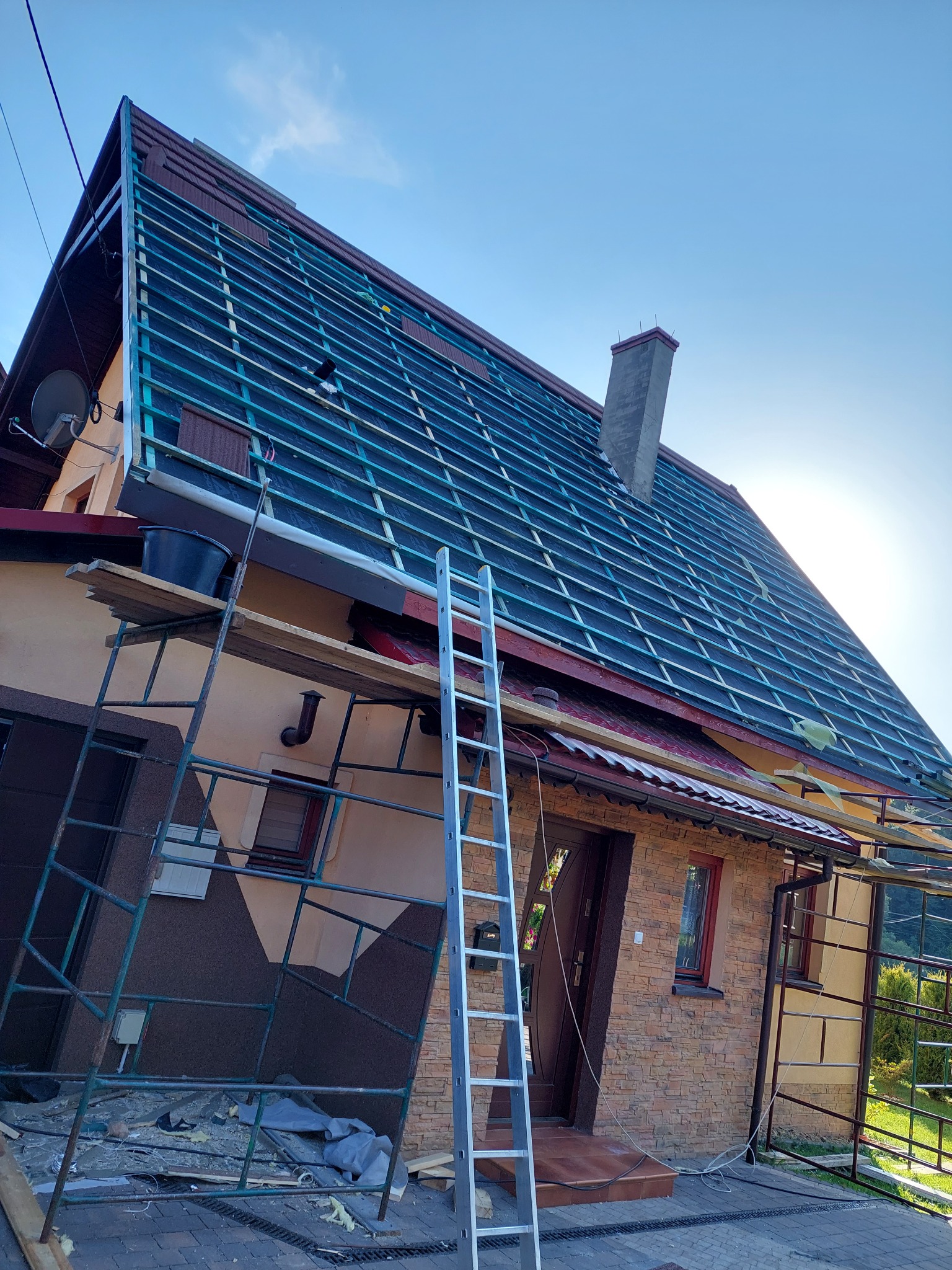 Dom w trakcie prac dekarskich: widoczne rusztowania, drabina oparta o ścianę, częściowo ułożona konstrukcja dachu z łatami i membraną, komin. Słoneczny dzień.