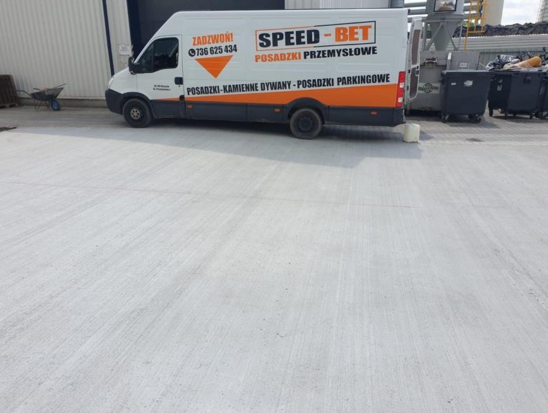 Świeżo wylana, jasnoszara betonowa posadzka przemysłowa przed budynkiem magazynowym, z zaparkowanym samochodem dostawczym firmy Speed-Bet w tle.