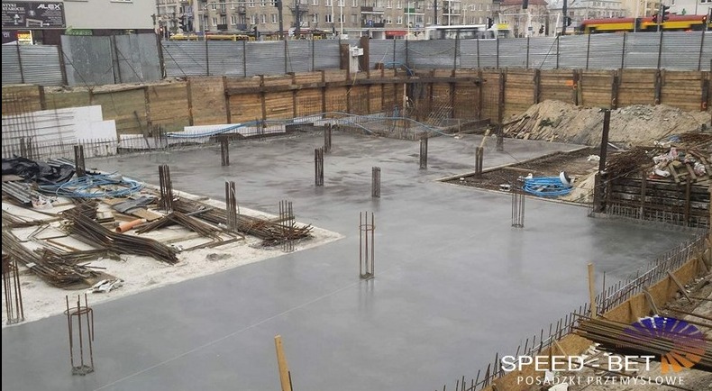 Świeżo wylana, gładka, szara posadzka na placu budowy, z widocznymi wystającymi prętami zbrojeniowymi i drewnianym szalunkiem. W tle widać budynki miejskie i przejeżdżający tramwaj.