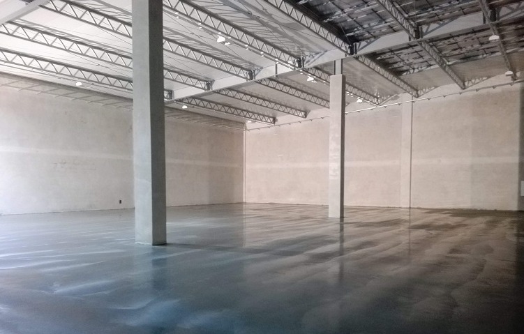 Wnętrze hali produkcyjnej z szarą, błyszczącą posadzką i betonowymi słupami nośnymi pod metalową konstrukcją dachu.