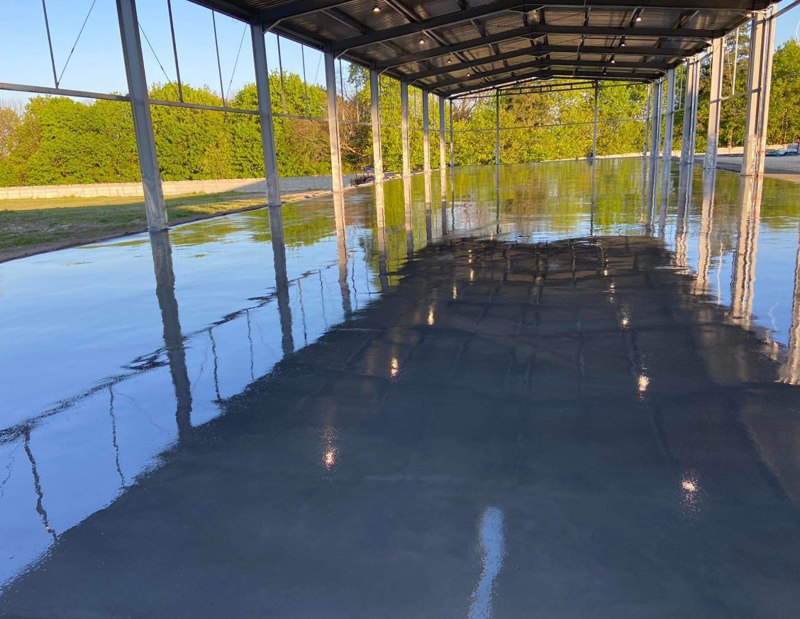 Błyszcząca, gładka, szara posadzka w hali magazynowej z metalową konstrukcją dachu, odbijająca światło lamp i elementy konstrukcji. Na zewnątrz widoczna zieleń drzew.