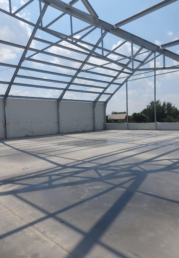 Wnętrze hali produkcyjnej w trakcie budowy, widoczna stalowa konstrukcja dachu rzucająca cienie na szarą, gładką posadzkę przemysłową, białe ściany boczne i fragment budynku na zewnątrz.