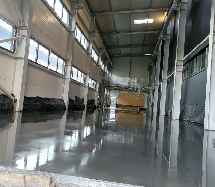 Świeżo wylana, błyszcząca, szara posadzka w hali przemysłowej z widocznymi oknami i stalową konstrukcją nośną.