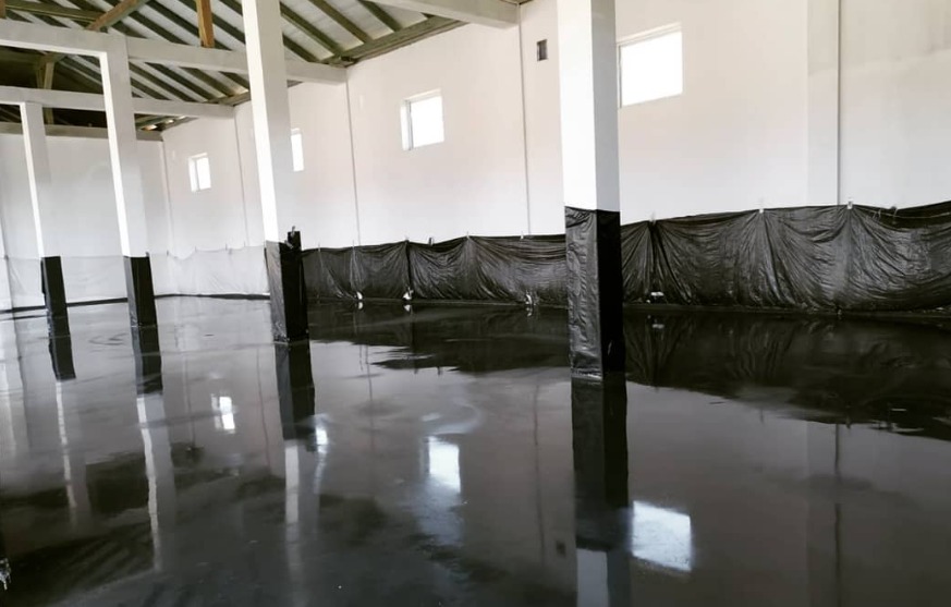 Świeżo wylana, błyszcząca, czarna posadzka w hali przemysłowej, zabezpieczona folią przy ścianach i kolumnach.