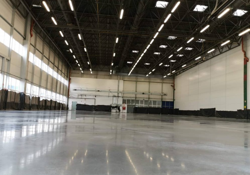 Błyszcząca, szara, gładka podłoga w hali przemysłowej z widoczną konstrukcją dachu i oświetleniem.