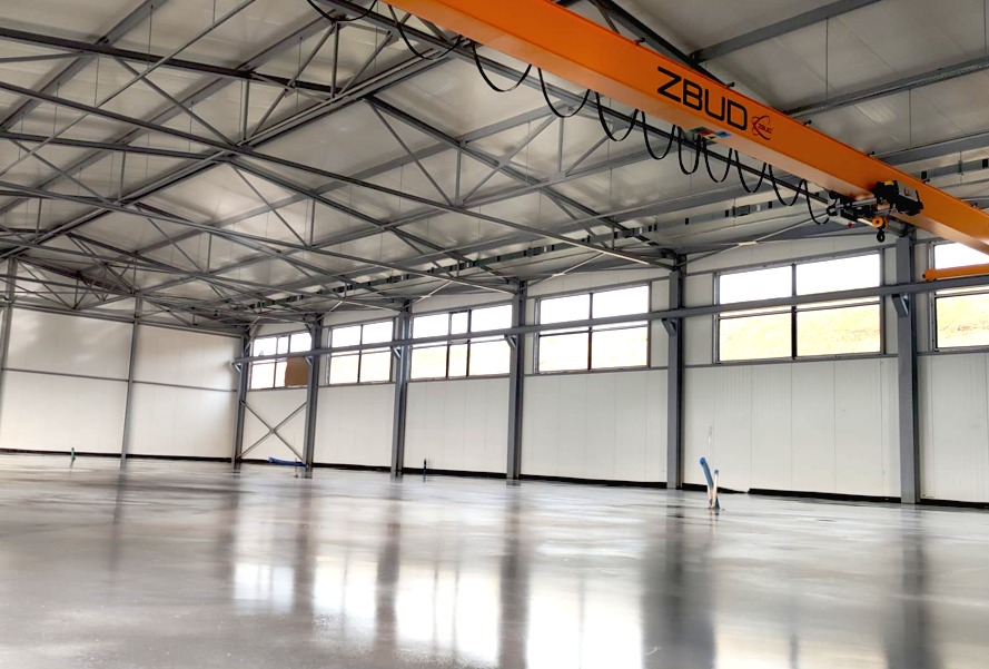 Świeżo wylana, błyszcząca posadzka w hali przemysłowej z widoczną suwnicą ZBUD pod sufitem i licznymi oknami wzdłuż ściany.