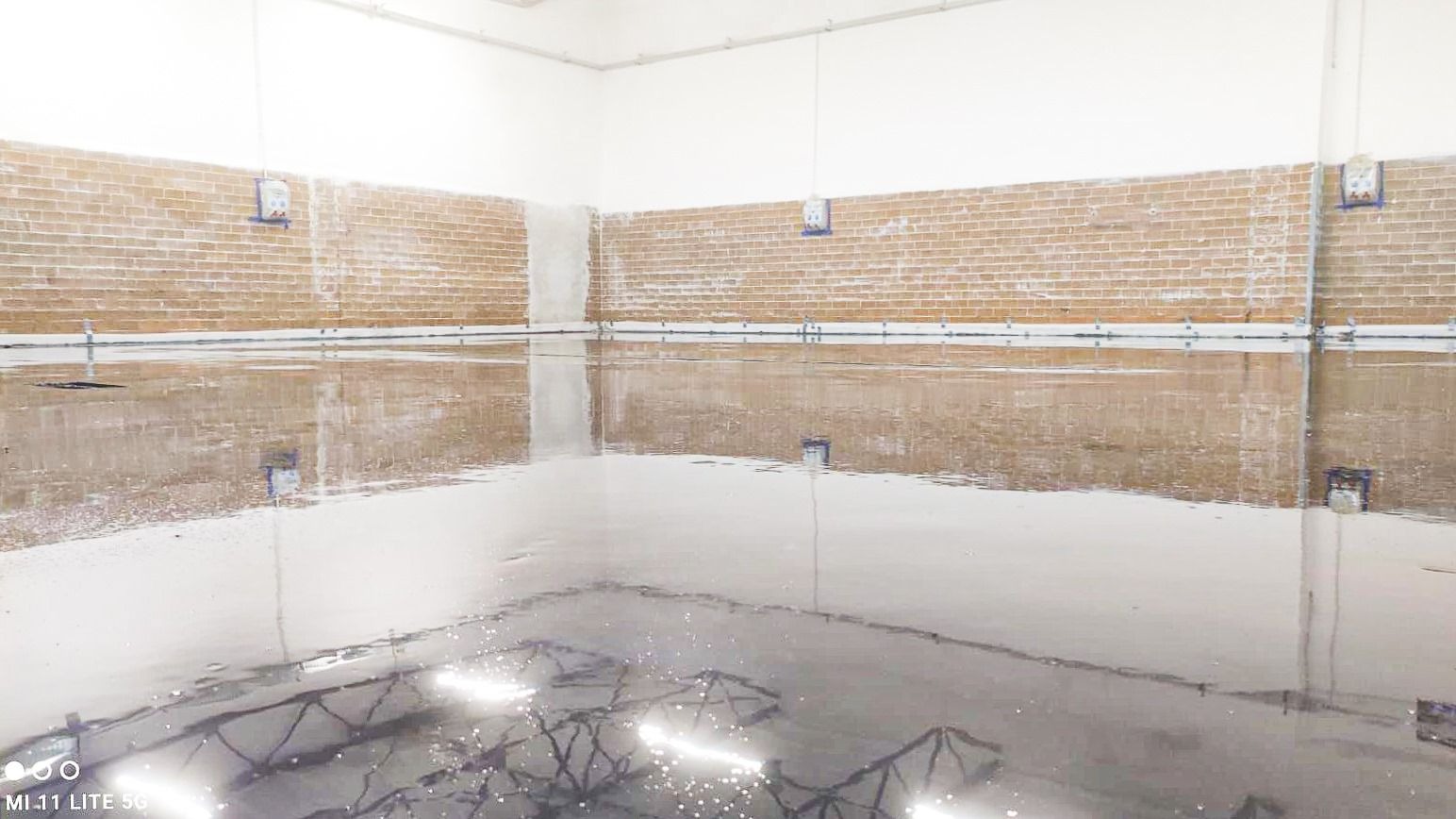 Świeżo wylana, mokra posadzka przemysłowa w hali, odbijająca ceglaną ścianę i oświetlenie sufitowe, z widocznymi elementami konstrukcyjnymi na ścianach.