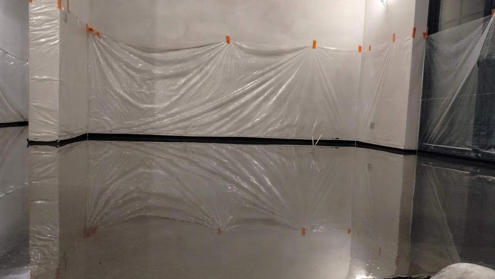 Świeżo wylana, błyszcząca posadzka przemysłowa zabezpieczona folią malarską przyklejoną pomarańczową taśmą, widoczne odbicia w mokrej powierzchni.