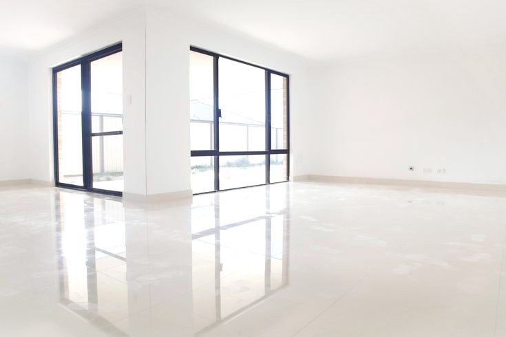 Białe, lśniące płytki podłogowe w pustym, jasnym pomieszczeniu z dużymi oknami i przesuwanymi drzwiami balkonowymi.