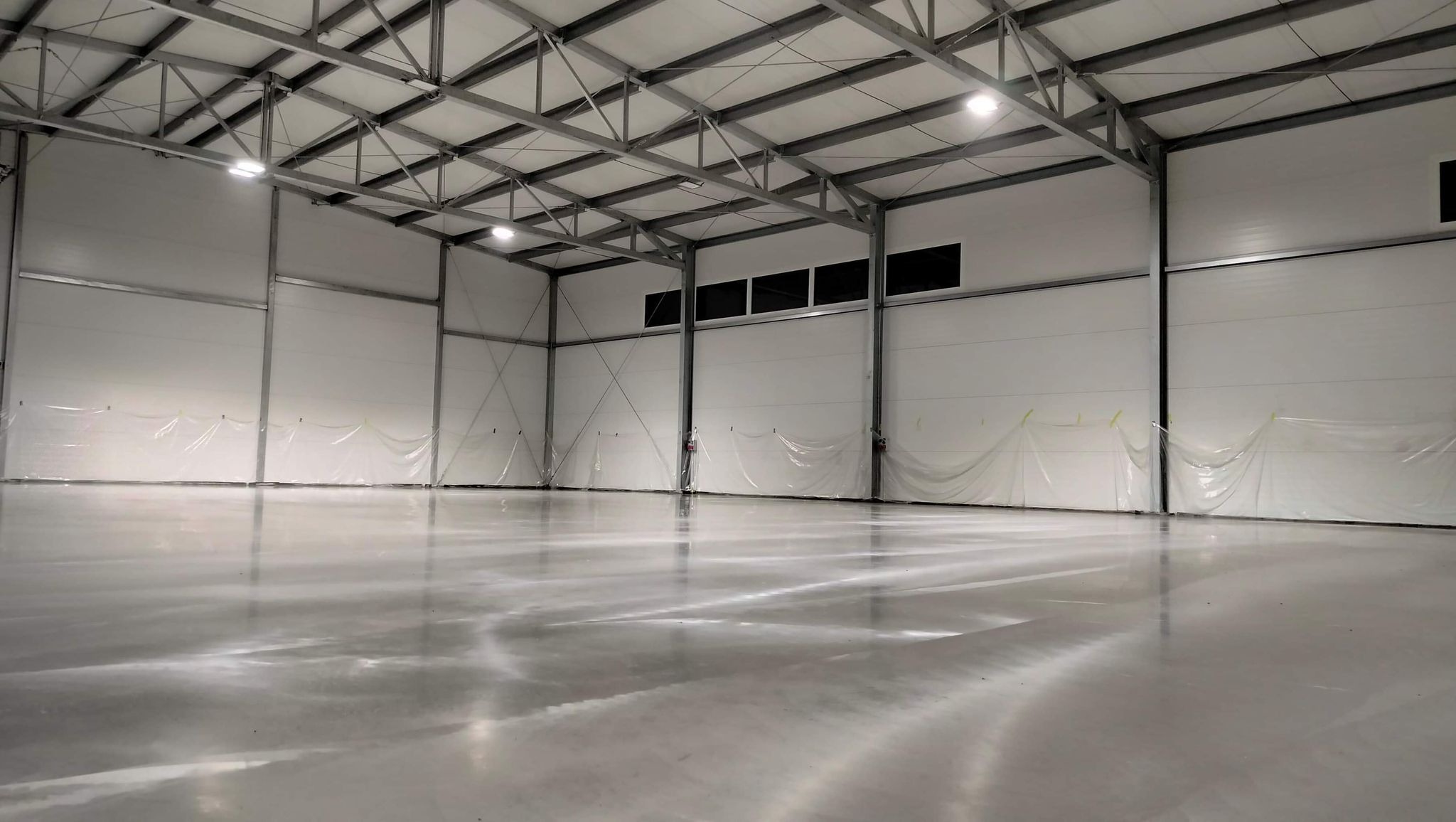 Błyszcząca, jasnoszara podłoga w hali przemysłowej, zabezpieczona folią ochronną przy ścianach z paneli, widoczne metalowe elementy konstrukcyjne dachu i oświetlenie.