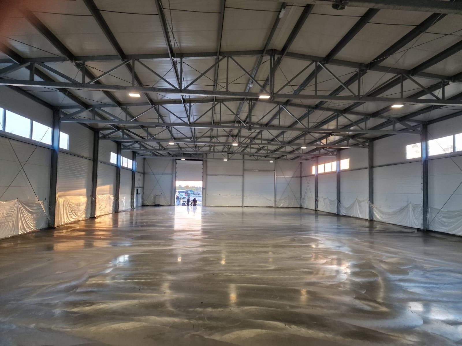 Wnętrze hali przemysłowej z nową, błyszczącą, betonową podłogą. Widoczne stalowe elementy konstrukcyjne dachu i ścian, okna w górnej części ścian oraz częściowo zasłonięte ściany folią ochronną.