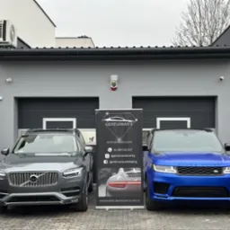 Dwa samochody, szary Volvo XC90 i niebieski Range Rover, prezentowane przed budynkiem z logo Gentleman's Auto Detailing & Spa oraz banerami reklamującymi ceramiczną ochronę Ultracoat.