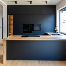 Kuchnie szafy Kita - Nowoczesna kuchnia z czarnymi szafkami i drewnianym blatem wyspy, oświetlona punktowymi lampami sufitowymi, widok na drewniane schody w tle.