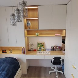 Kuchnie szafy Kita - Pokój dziecięcy z zabudową meblową: białe szafy z drewnianymi wstawkami i oświetleniem LED, biurko z krzesłem na kółkach, łóżko z niebieską narzutą, wiszące lampy ażurowe.