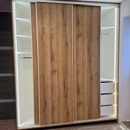 Kuchnie szafy Kita - Nowoczesna szafa przesuwna z drewnopodobnymi frontami i białym wnętrzem, oświetlona taśmami LED, z widocznymi półkami, drążkiem i szufladami, na tle paneli podłogowych.