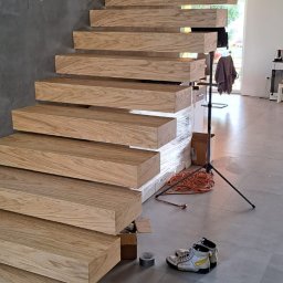 Kuchnie szafy Kita - Montaż drewnianych schodów wiszących z jasnego drewna, widoczne buty robocze, taśma malarska i kable na szarych płytkach podłogowych, surowy beton na ścianie.