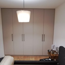 Kuchnie szafy Kita - Beżowa szafa wnękowa z długimi, czarnymi uchwytami, widoczna z perspektywy łóżka w sypialni, z lampą wiszącą nad szafką nocną i częścią otwartych drzwi po lewej stronie.