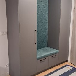 Kuchnie szafy Kita - Szara szafa wnękowa z siedziskiem i tapicerowanym panelem w kolorze morskim, widoczne zabezpieczenia podłogi podczas montażu.