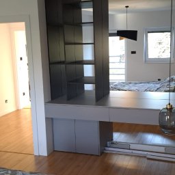 Kuchnie szafy Kita - Szara zabudowa meblowa z półkami i szafkami, pełniąca funkcję ścianki działowej w sypialni, z widocznym łóżkiem w tle i drewnianą podłogą.