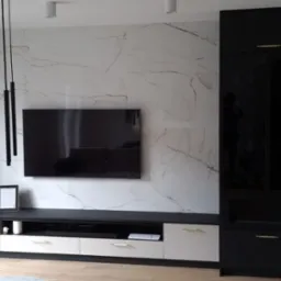 Nowoczesny salon z czarno-białymi meblami na wymiar, marmurową ścianą i wiszącymi lampami tubami, z widokiem na drewniane schody w tle.