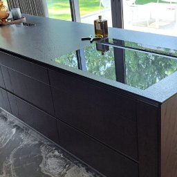 Kuchnie szafy Kita - Wyspa kuchenna z ciemnego drewna z blatem o fakturze kamienia, wbudowaną płytą indukcyjną i szufladami, uzupełniona przezroczystą butelką i dekoracjami, otoczona krzesłami barowymi i dużymi oknami...