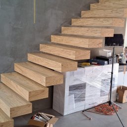 Kuchnie szafy Kita - Drewniane schody wspornikowe z jasnego drewna dębowego zamontowane na szarej ścianie, widoczne przygotowania do montażu, meble zabezpieczone folią stretch, karton z silikonami montażowymi na podłodze.