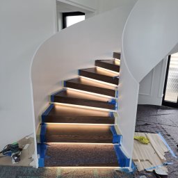 Kuchnie szafy Kita - Drewniane schody spiralne z podświetleniem LED w trakcie montażu, zabezpieczone niebieską taśmą malarską, widoczne narzędzia i materiały budowlane.