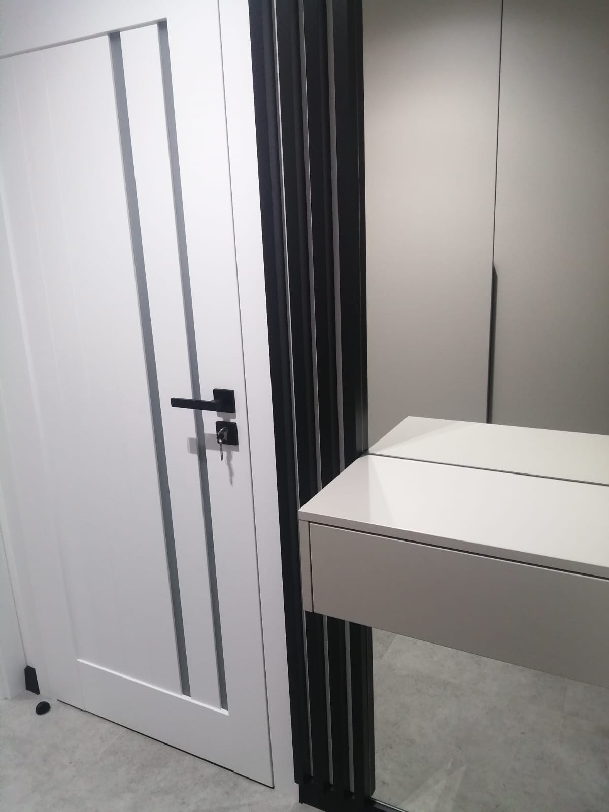 Białe drzwi wewnętrzne z czarną klamką i kluczem, obok nowoczesna, jasna szafa z lustrem i czarnymi, pionowymi listwami dekoracyjnymi.