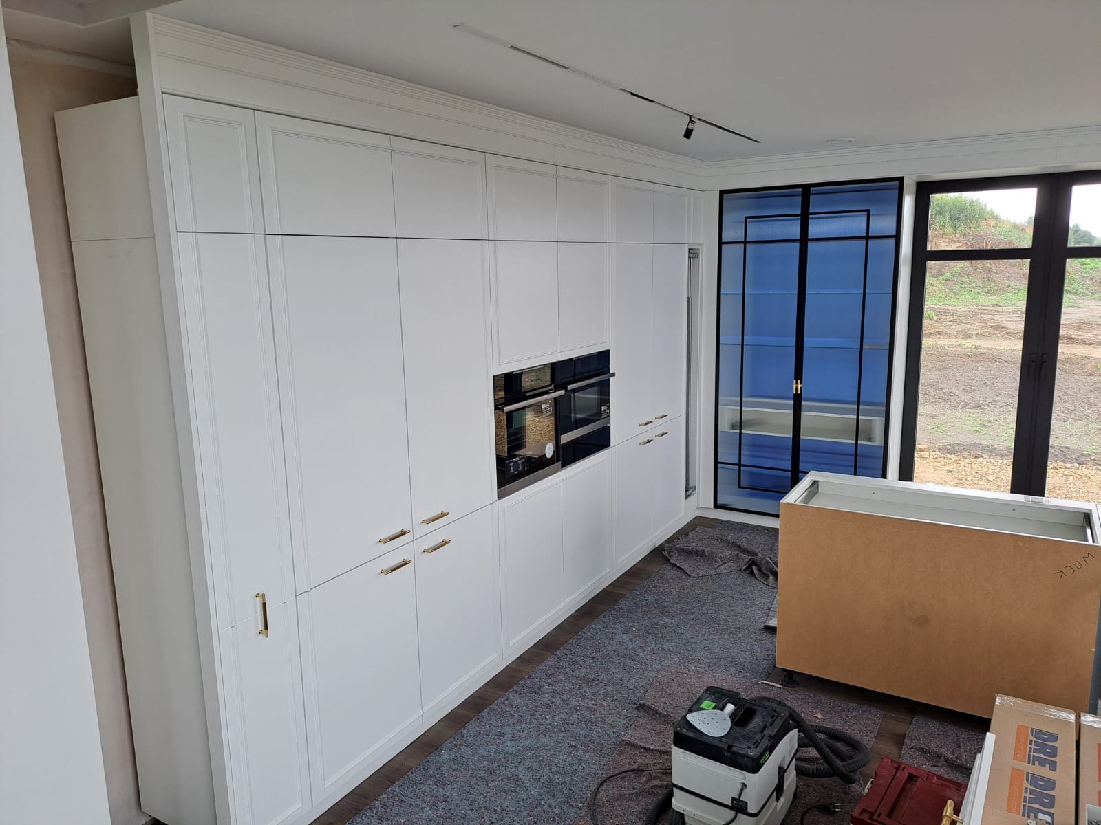 Wysoka zabudowa kuchenna w kolorze białym z piekarnikiem i szafkami, widoczne elementy montażowe i narzędzia, okno z czarną ramą w tle.