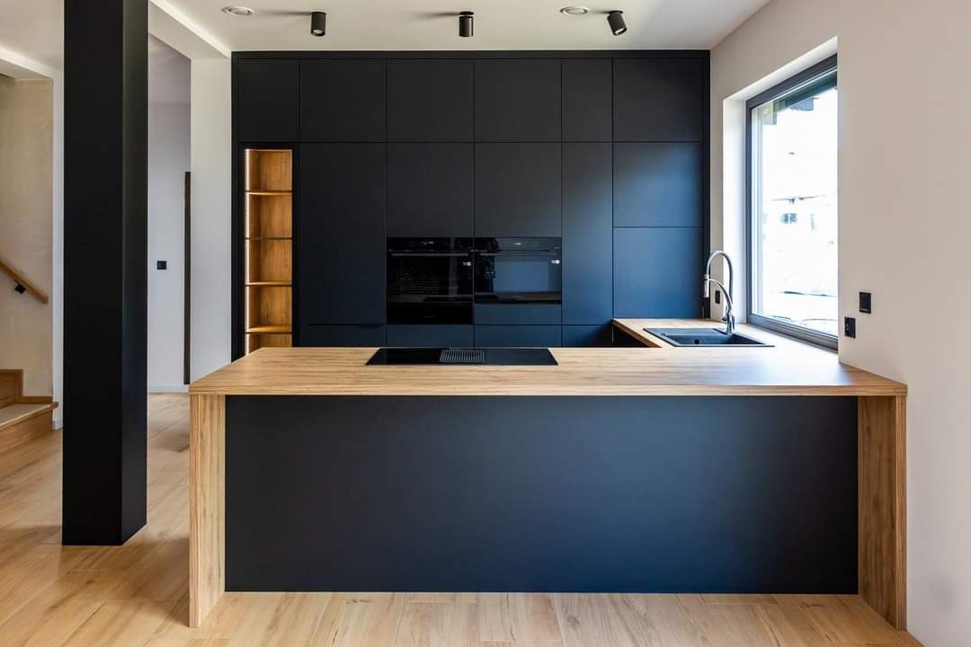 Nowoczesna kuchnia z czarnymi szafkami i drewnianym blatem wyspy, oświetlona punktowymi lampami sufitowymi, widok na drewniane schody w tle.