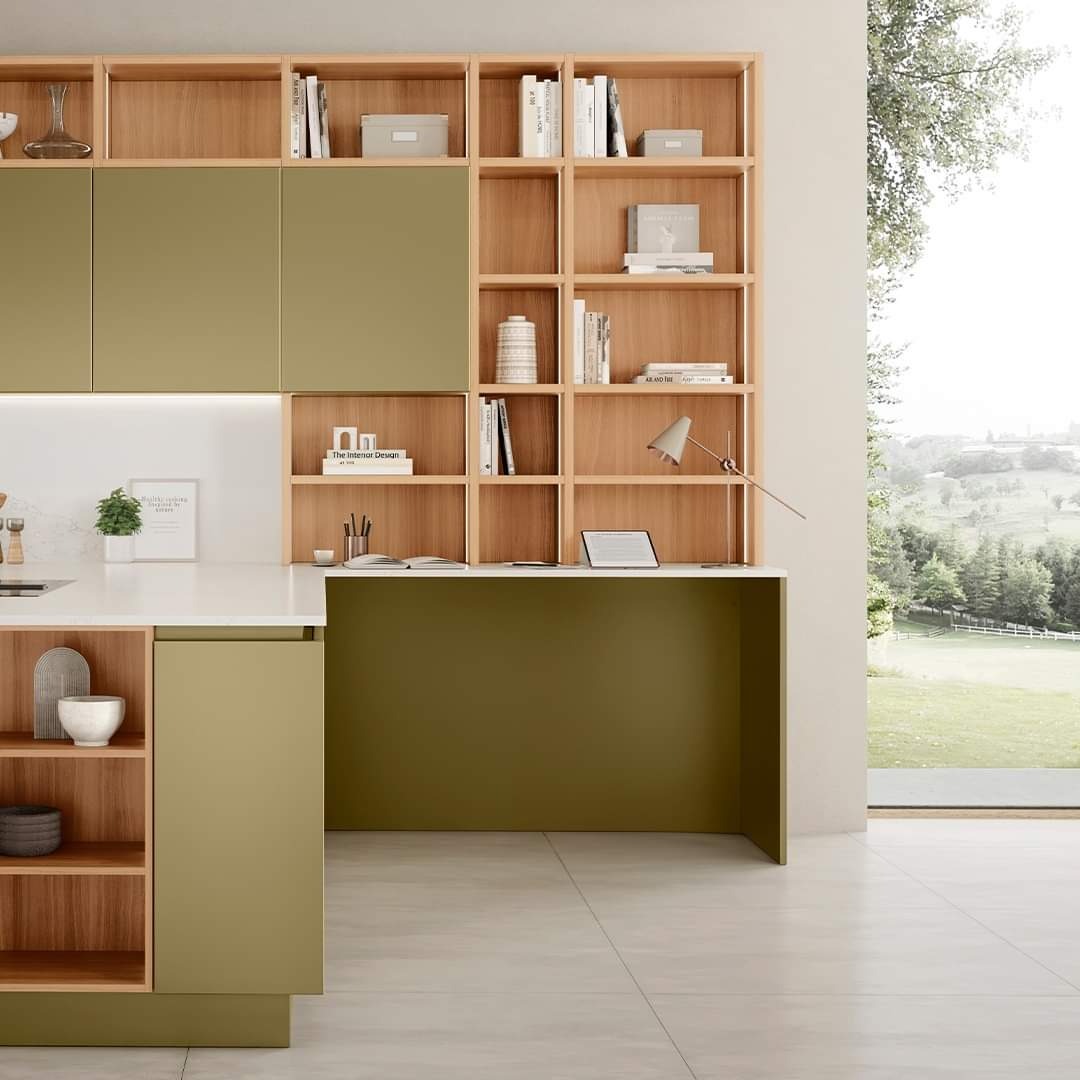 Nowoczesna kuchnia z zabudową w odcieniach oliwkowej zieleni i naturalnego drewna, z otwartymi półkami i blatem roboczym przechodzącym w biurko, z widokiem na zieleń za oknem.