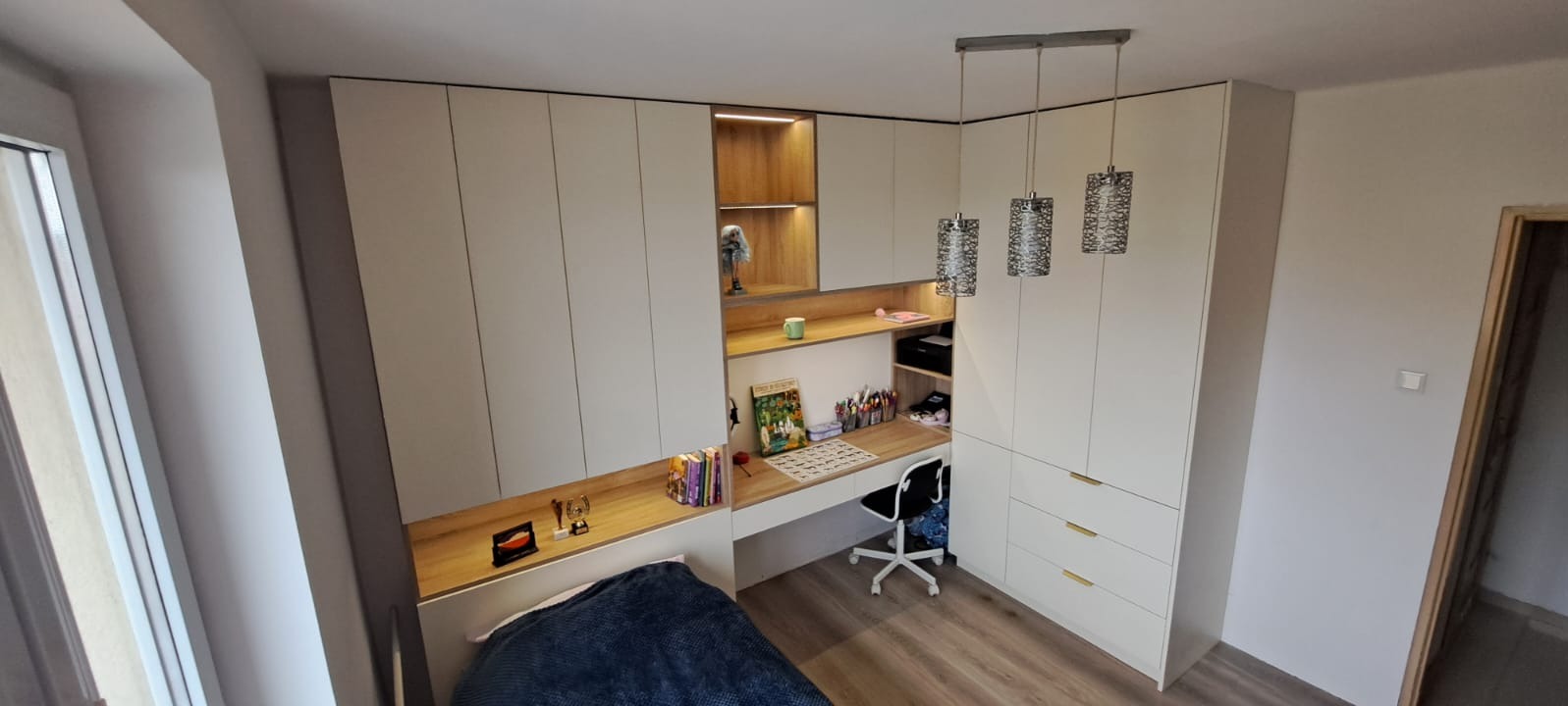 Pokój młodzieżowy z zabudową meblową: białe szafy z drewnianym biurkiem i półkami, oświetlenie LED w półkach, wiszące lampy dekoracyjne, łóżko z niebieską narzutą, widok z góry.