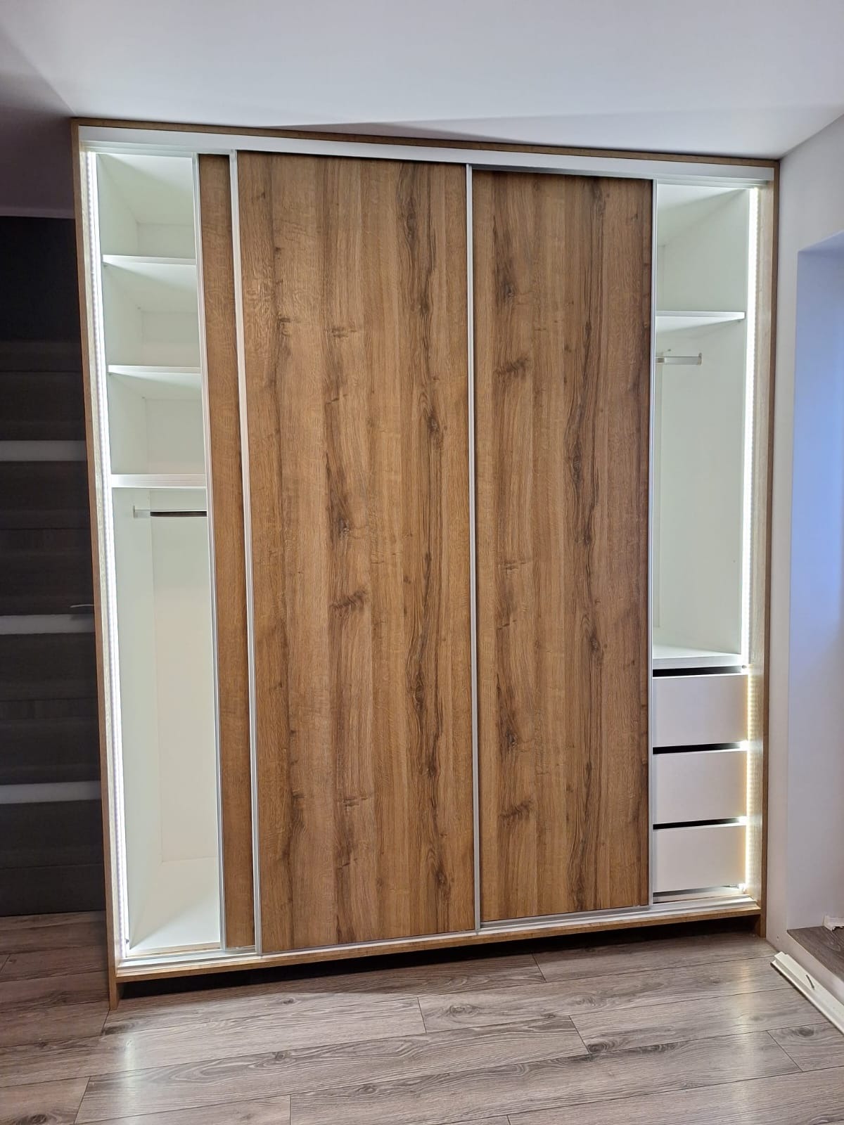 Nowoczesna szafa przesuwna z drewnopodobnymi frontami i białym wnętrzem, oświetlona taśmami LED, z widocznymi półkami, drążkiem i szufladami, na tle paneli podłogowych.