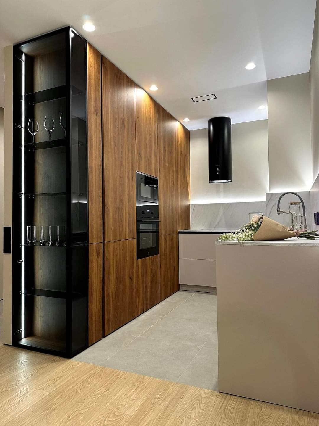Nowoczesna kuchnia z drewnianymi frontami, czarnym okapem cylindrycznym i przeszkloną witryną z podświetleniem LED, uzupełniona jasnym blatem i bukietem kwiatów.