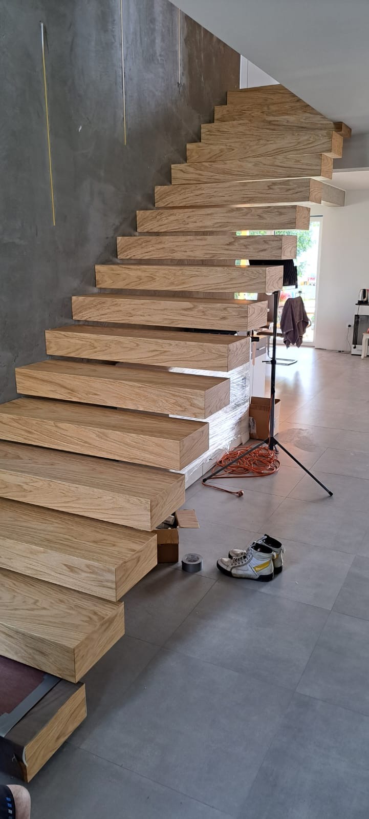 Montaż drewnianych schodów wiszących z jasnego drewna, widoczne buty robocze, taśma malarska i kable na szarych płytkach podłogowych, surowy beton na ścianie.
