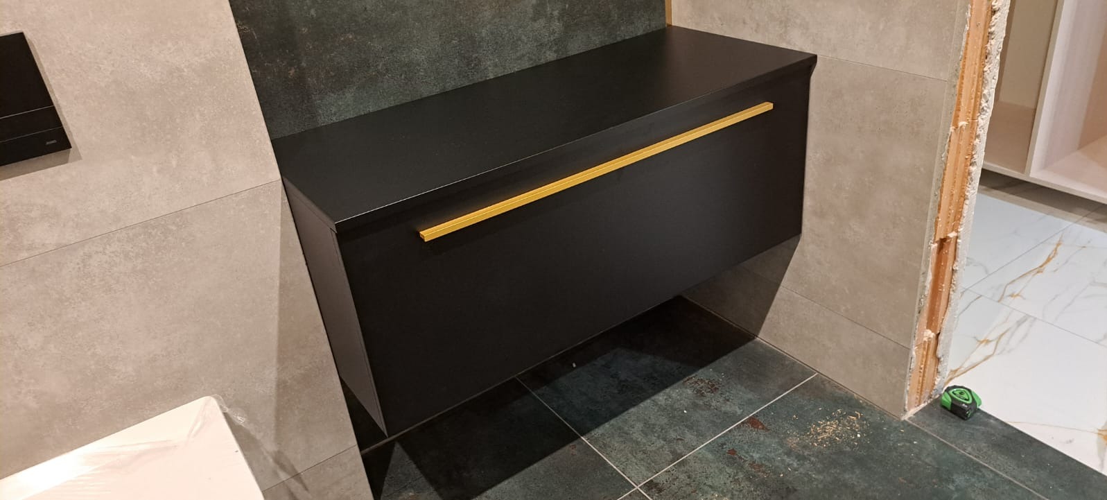 Czarna szafka łazienkowa wisząca z złotym uchwytem, zamontowana na tle szarych i zielonych płytek, widoczny fragment toalety oraz niedokończona zabudowa w tle.