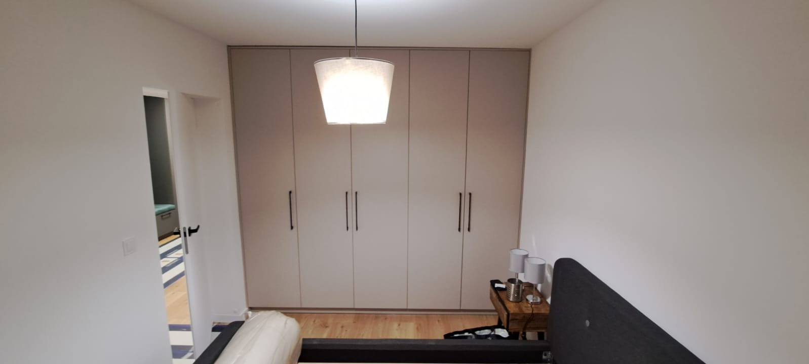 Beżowa szafa wnękowa z długimi, czarnymi uchwytami, widoczna z perspektywy łóżka w sypialni, z lampą wiszącą nad szafką nocną i częścią otwartych drzwi po lewej stronie.