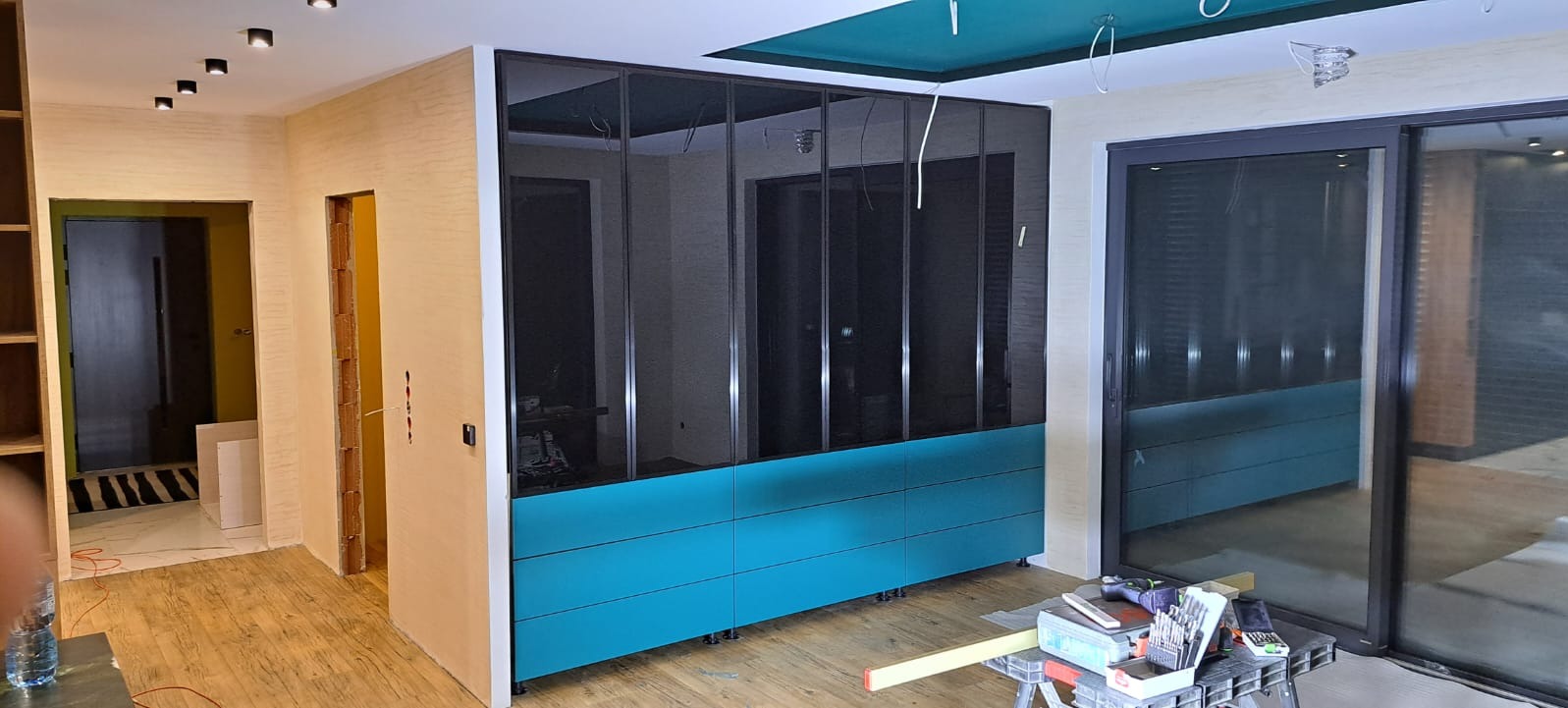 Nowoczesna szafa wnękowa z czarnymi, lustrzanymi drzwiami i turkusowymi szufladami, widoczna w trakcie montażu, z narzędziami na pierwszym planie i surowym wykończeniem wnętrza.