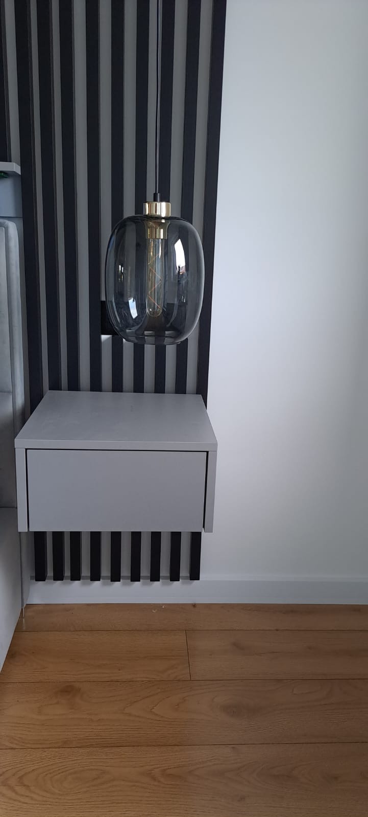Szara szafka nocna wisząca na ścianie z czarnymi, pionowymi listwami dekoracyjnymi pod spodem, oświetlona szklaną lampą wiszącą z żarówką edisona na tle ściany z pionowymi panelami dekoracyjnymi...