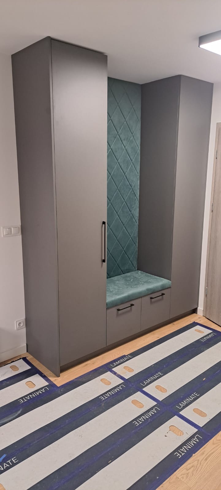 Szara szafa wnękowa z siedziskiem i tapicerowanym panelem w kolorze morskim, widoczne zabezpieczenia podłogi podczas montażu.