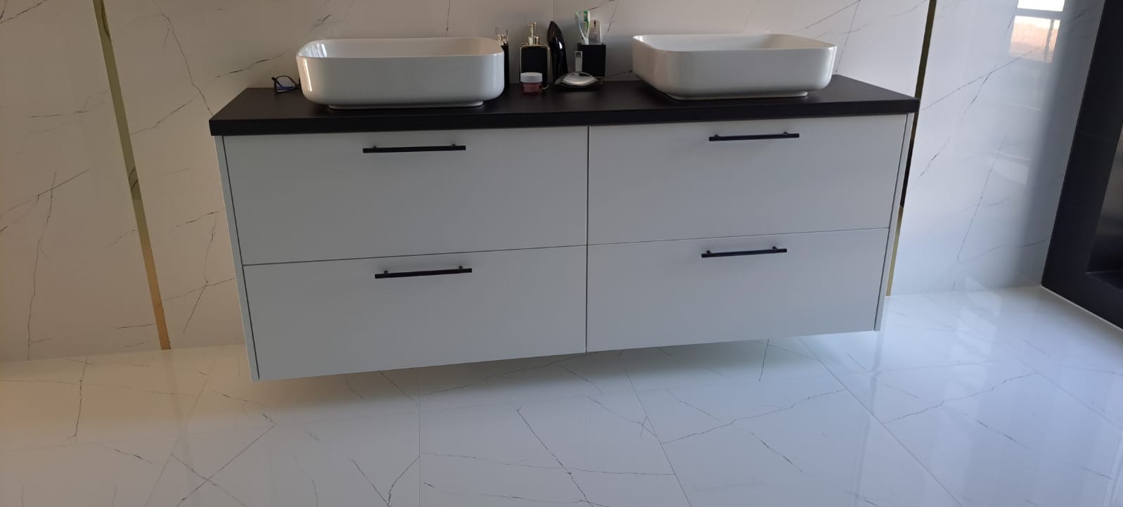 Nowoczesna szafka łazienkowa w kolorze białym z czarnym blatem i dwiema umywalkami nablatowymi, minimalistyczne czarne uchwyty, białe płytki podłogowe i ścienne z szarym wzorem żyłek.